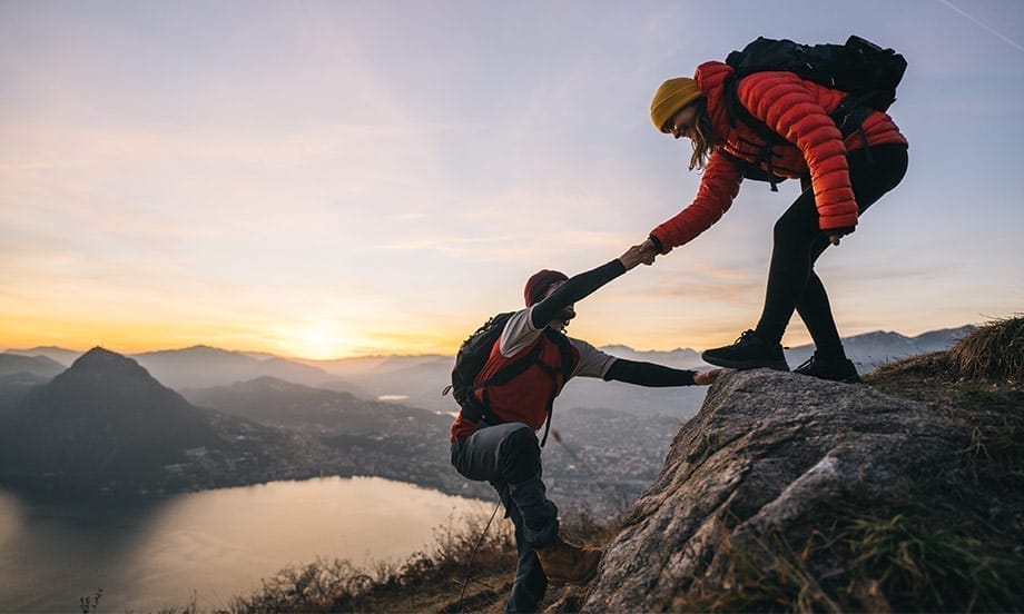 Frau auf einem Felsen reicht ihrem Partner die Hand und zieht ihn nach oben